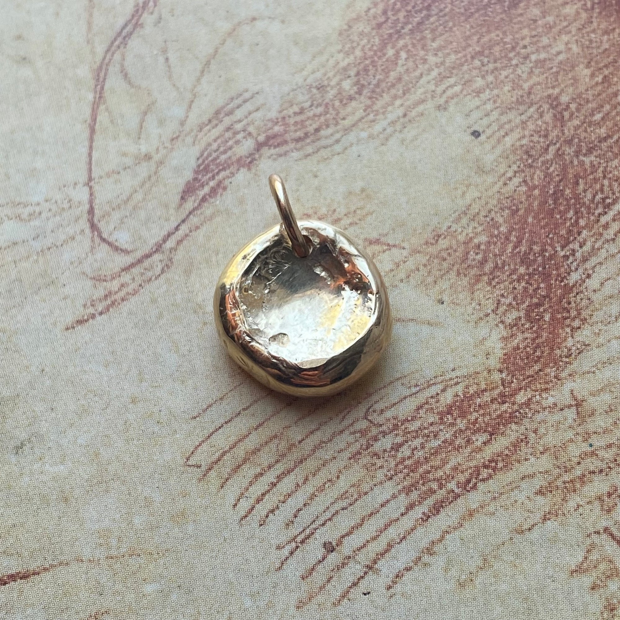 A small polished gold pendant with a hammered, organic textured surface and a top loop, resting on a vintage-style beige paper background with reddish-brown sketch lines.