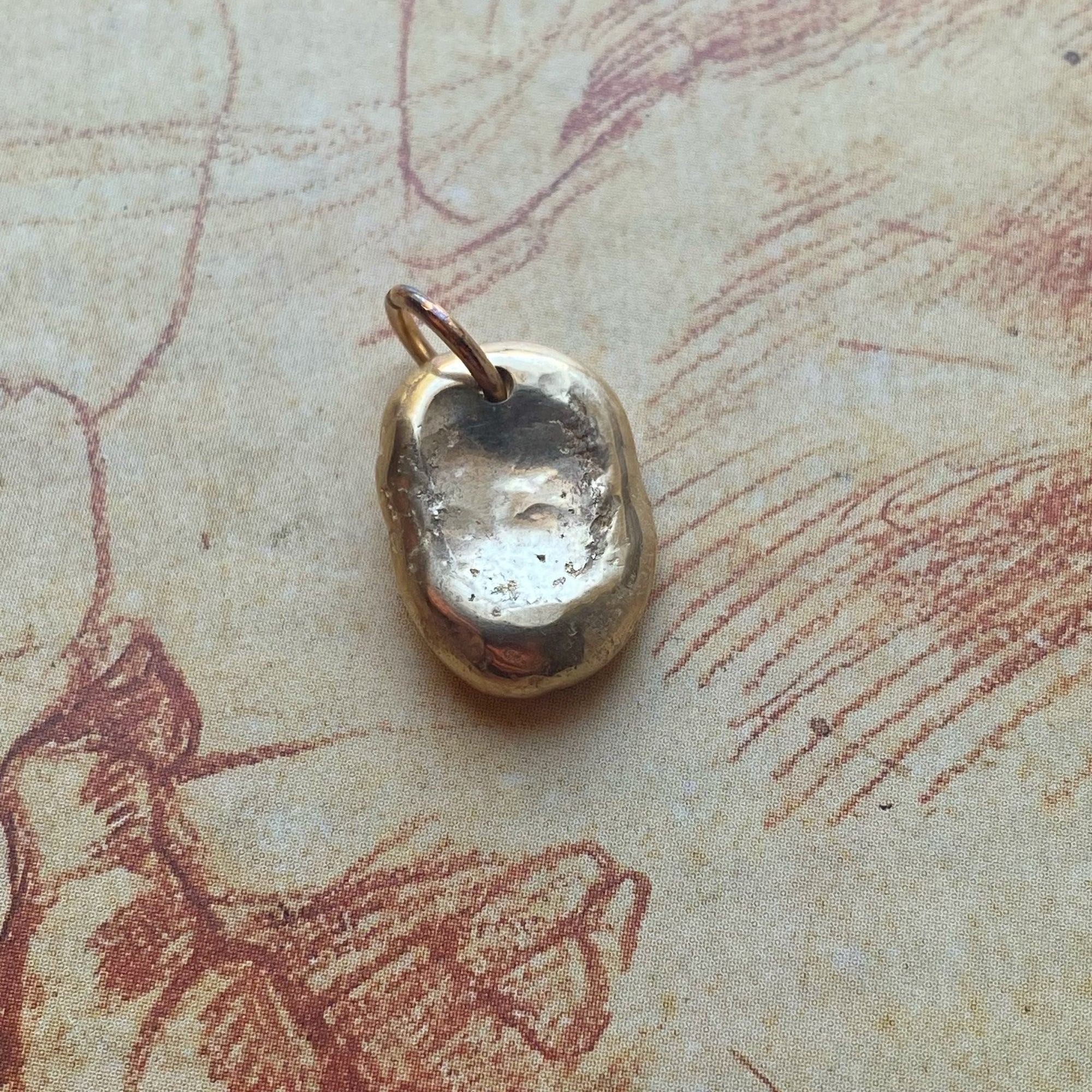 A small, organic-shaped gold charm with a textured, hammered surface and a loop at the top, resting on a vintage-style beige paper background with red sketches.
