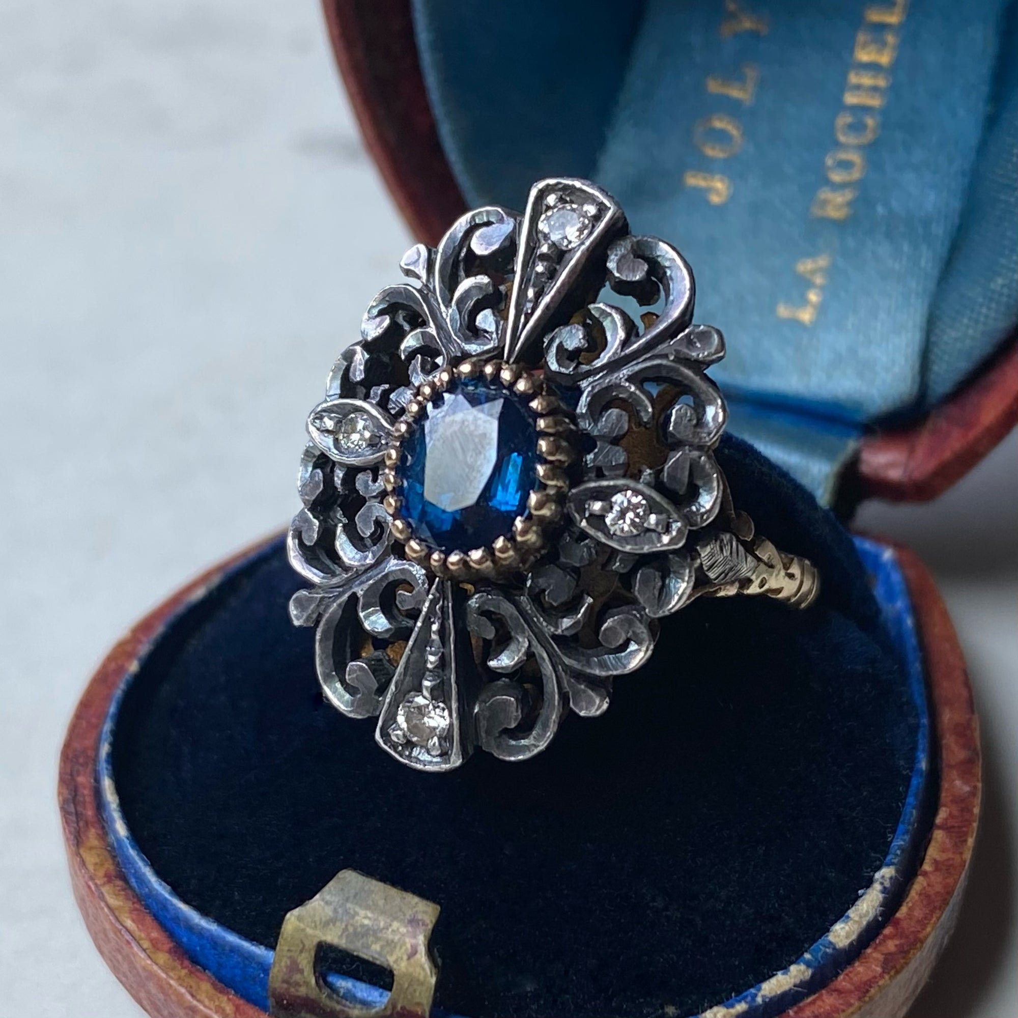 An ornate antique filigree ring with a blue oval gemstone and small clear stones, displayed inside an open blue velvet ring box.