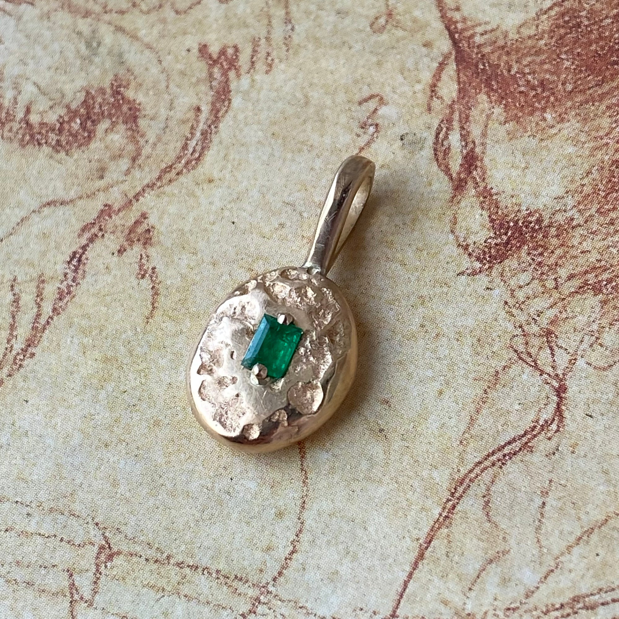 A textured gold pendant with a small rectangular green emerald-cut gemstone, displayed on a vintage-style beige map background.