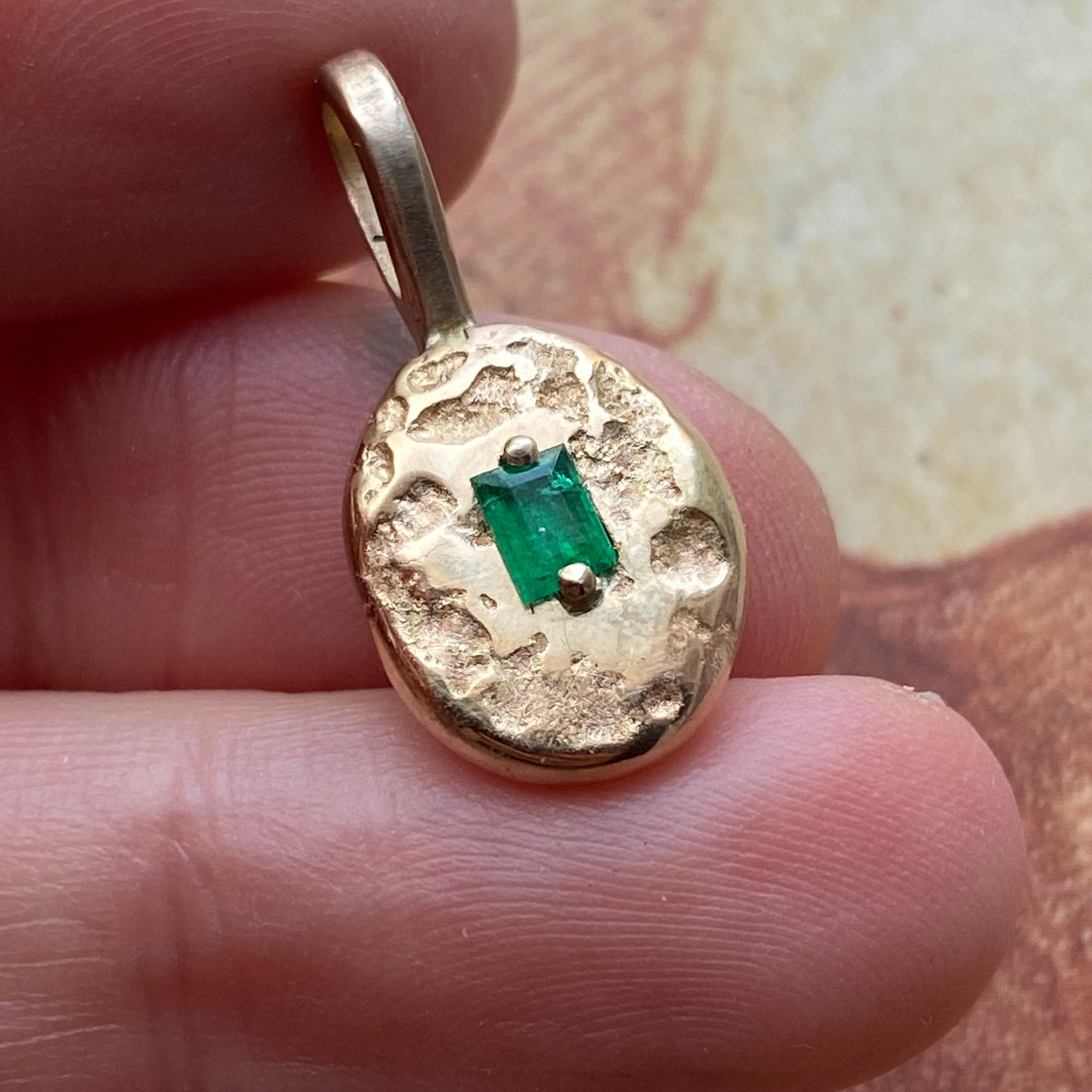 A gold pendant with a hammered texture and a small green rectangular gemstone in the center, held between fingers.