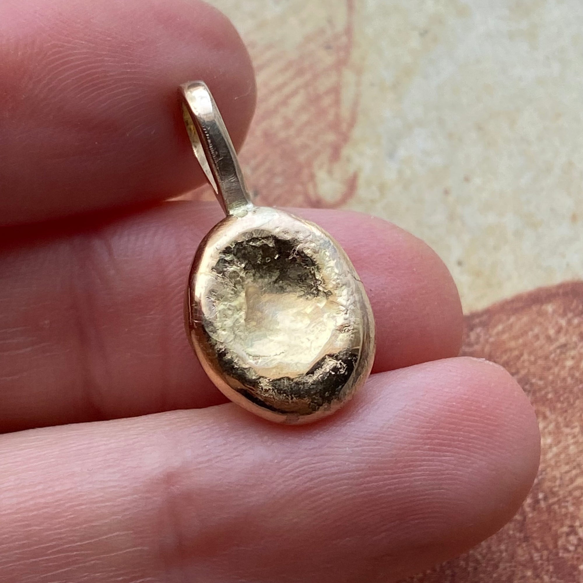 A small gold pendant with a highly textured, organic, hammered surface is held between a person's fingers against a neutral beige background.