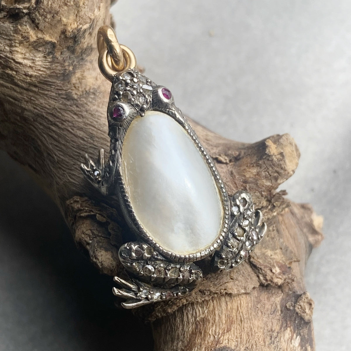 A frog-shaped pendant with a large oval natural pearl center, set in silver with small red gemstone eyes and diamond details, resting on a piece of driftwood.